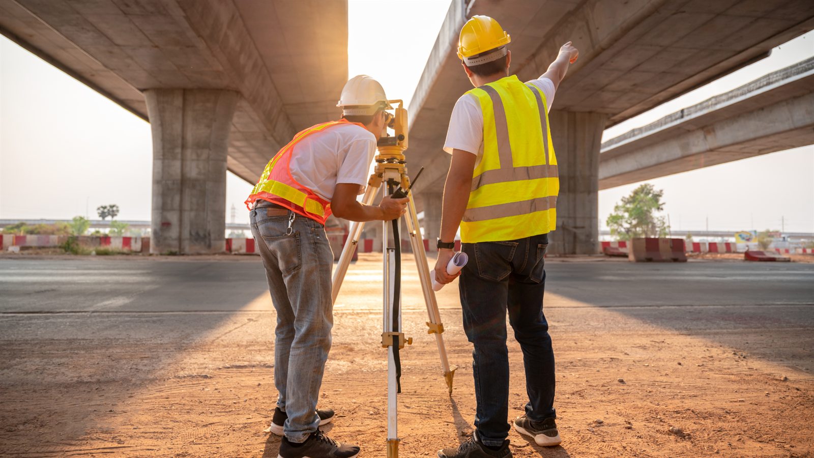 Dos ingenieros con casco y chaleco de seguridad revisan una obra vial con un nivel topográfico bajo un puente en construcción