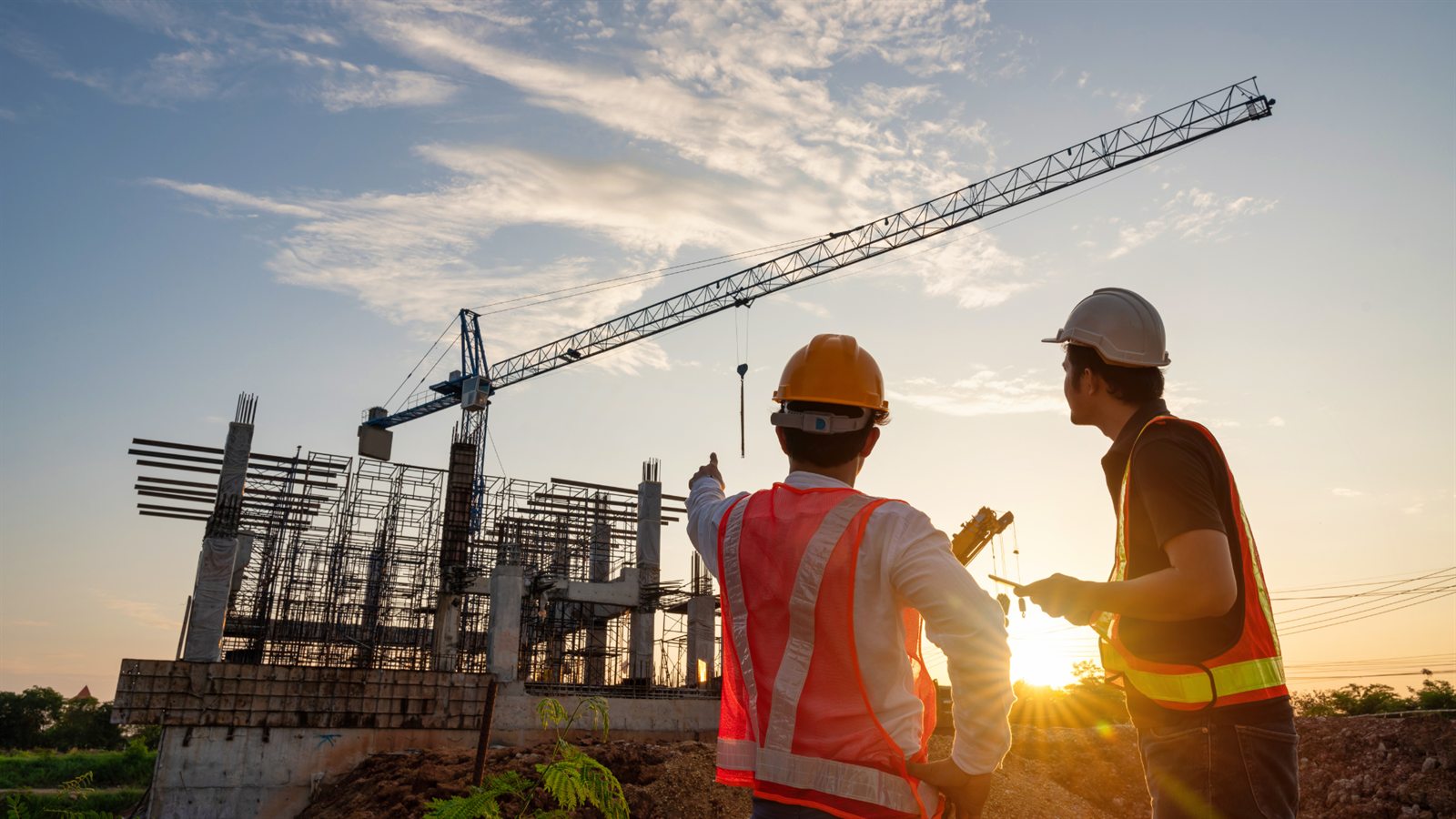 Dos ingenieros con casco y chaleco de seguridad revisan una obra en construcción con una grúa torre al atardecer
